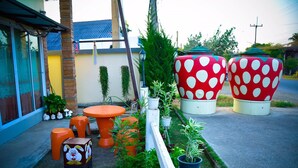 A garden area with a round orange table, orange stools, and a small decorative rabbit statue. There are large red and white polka-dotted planters and various potted plants. - Sabaidee Place (Phetchabun)