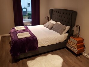 A neatly made bed with a purple blanket, white linens, and several pillows. There is a white fluffy rug on the floor, a bedside table with a lamp, and a small wooden nightstand with a patterned box. - First St John's Hill Apartment 2 (Edinburgh)