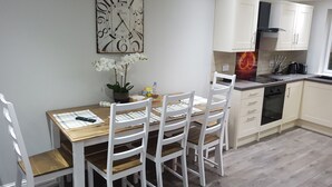 A modern kitchen with white cabinetry, a wooden dining table with four chairs, a potted orchid, and a wall clock. - First St John's Hill Apartment 2 (Edinburgh)