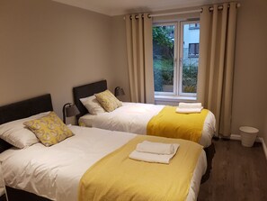 A hotel room with two beds, beige walls, a window with curtains, and a view of greenery outside. - First St John's Hill Apartment 2 (Edinburgh)
