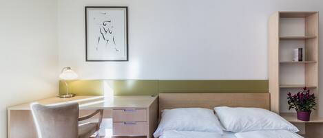 Quarto de hotel com uma cama, uma escrivaninha com cadeira, mesa de cabeceira, prateleira na parede e uma obra de arte emoldurada.