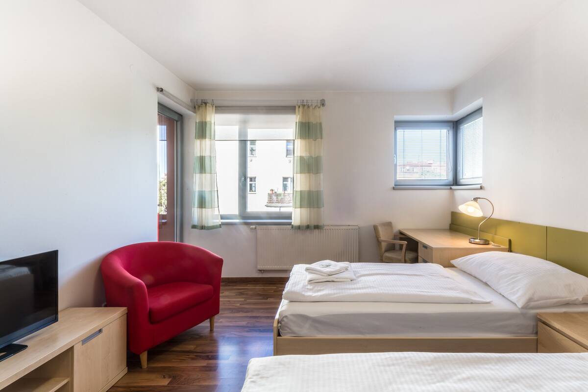 A hotel room with two beds, a TV, a red chair, a desk, and a window with curtains.