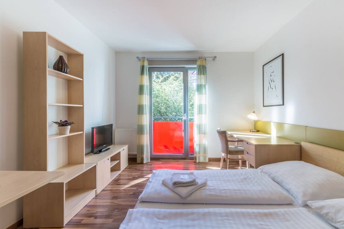 A hotel room with a bed, a desk, a TV, a bookshelf, and a view of the outside.