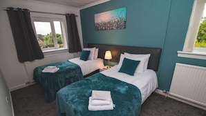 A hotel room with two beds, teal bedding, a nightstand with a lamp, and a painting on the wall. - Bentley Hotel (Motherwell)