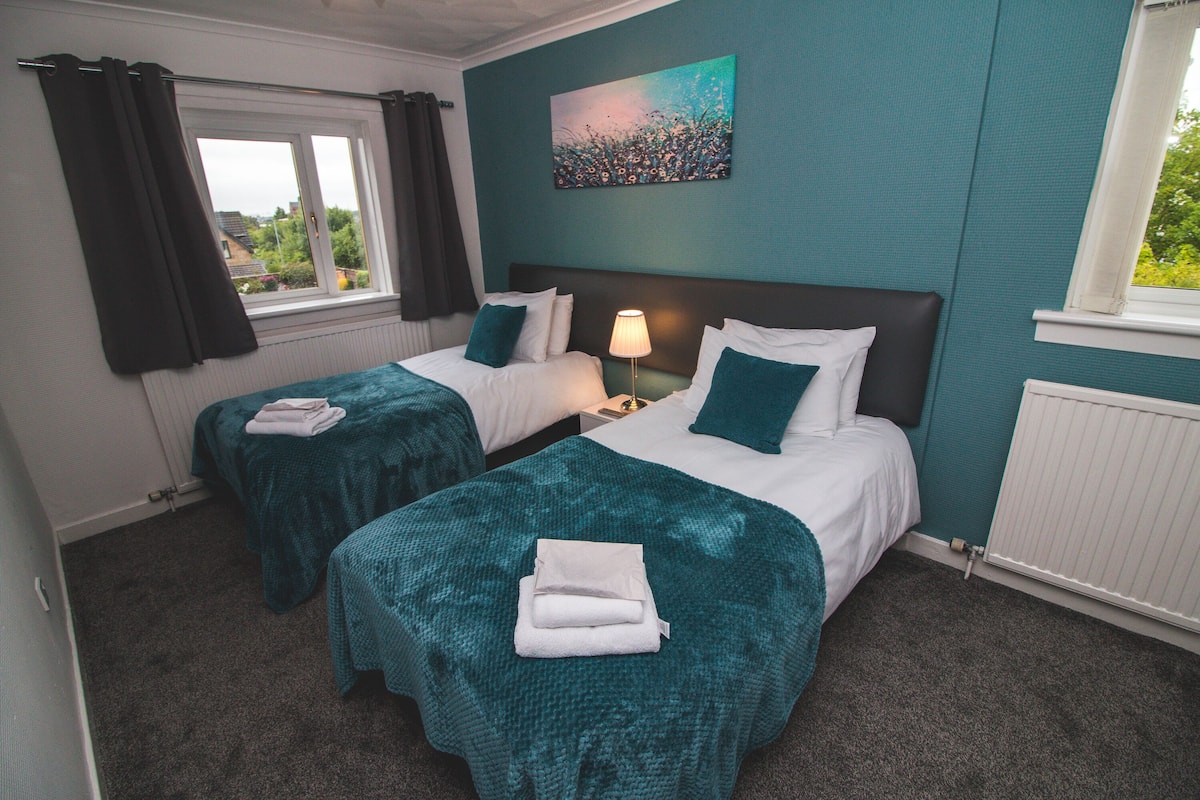 A hotel room with two beds, teal bedding, a nightstand with a lamp, and a painting on the wall.