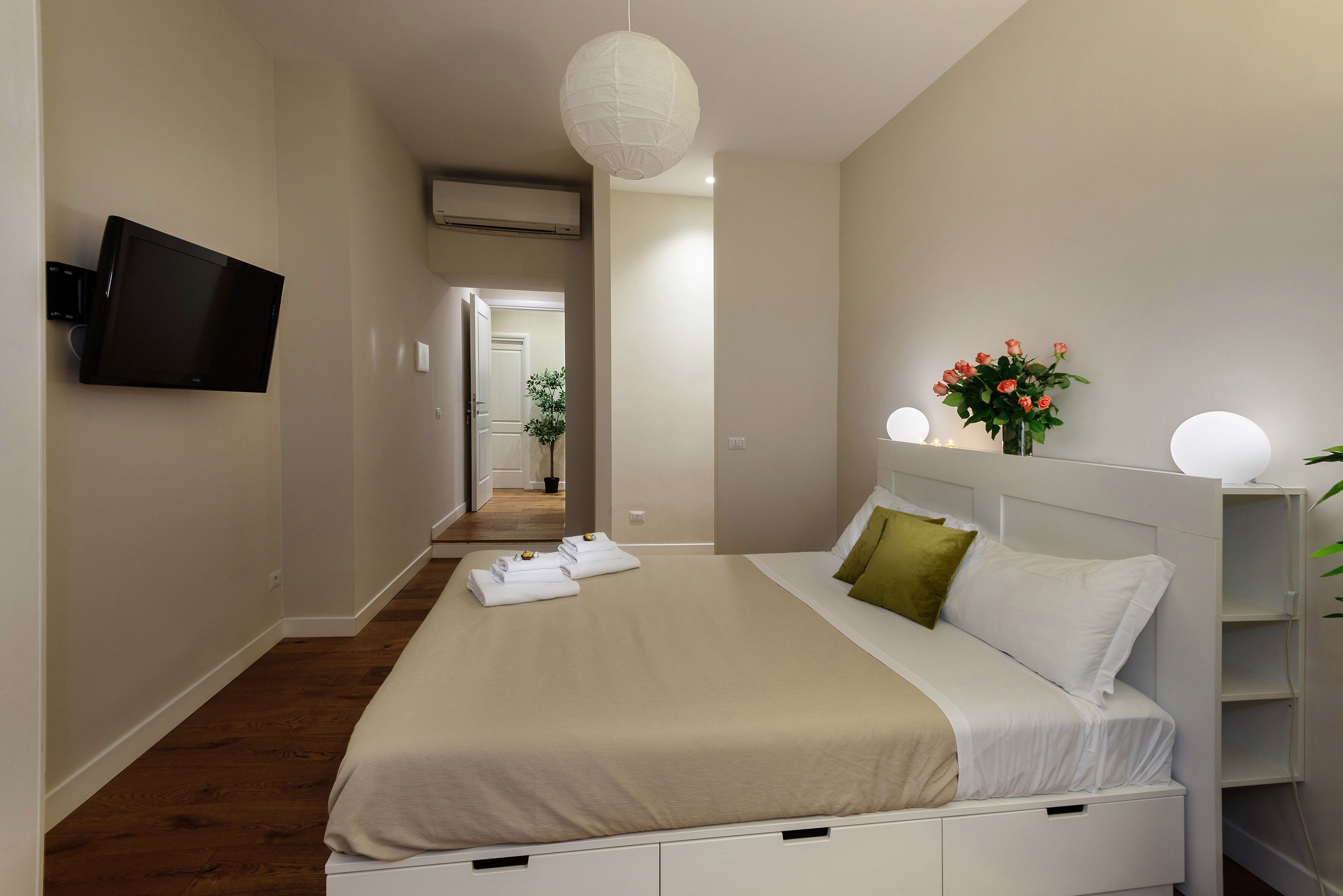 Een slaapkamer met een bed, een tv aan de muur, een nachtkastje met een lamp en een bloevas.