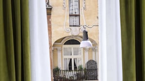 A view through a window with green curtains, revealing a building with a balcony, a hanging light fixture, and potted plants. - Tula Inn (Rome)