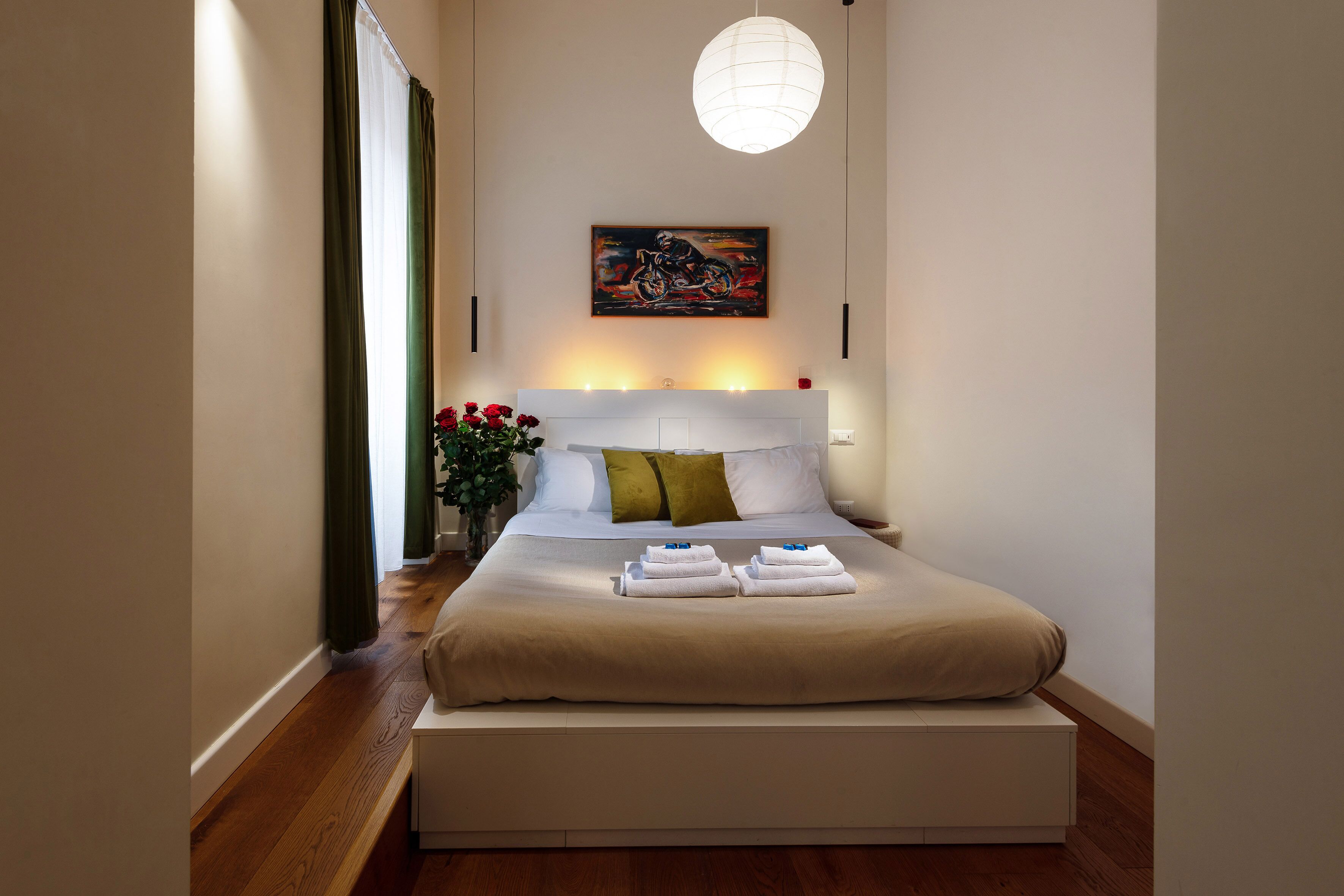 Una cama bien hecha con cabecera, una mesita de noche con un florero y un cuadro en la pared.