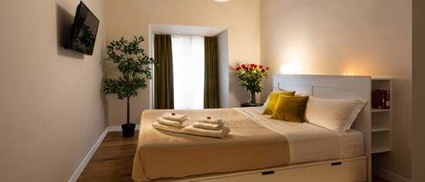 A modern hotel room with a large bed, a flat-screen TV mounted on the wall, a bedside table with a lamp, and a small potted plant.