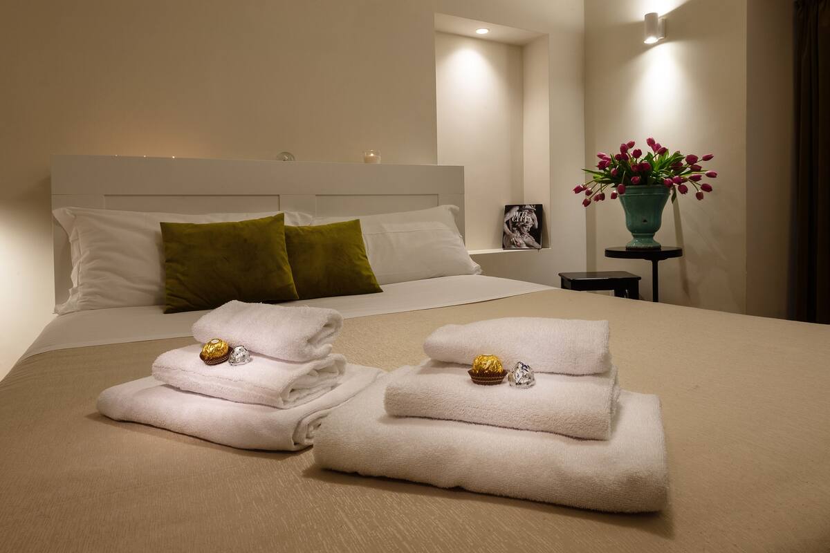 A neatly made bed with white and green pillows, a small bedside table with a picture frame, and a vase with flowers on a nightstand.
