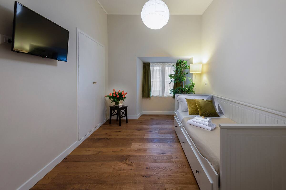 A room with a flat-screen TV mounted on the wall, a wooden floor, a bed with a white headboard, a small table with a vase of flowers, and a window with curtains.