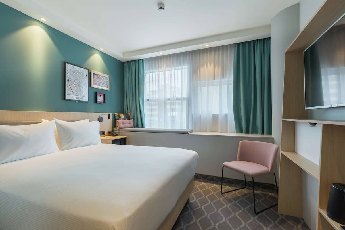 A hotel room with a large bed, a chair, a TV, a window with curtains, and artwork on the wall.