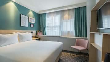 Habitación de hotel con una cama grande, una silla, televisión, ventana con cortinas y obras de arte en la pared.