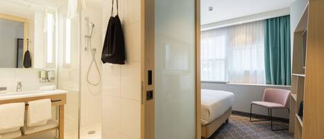 Una habitación de hotel moderna con cabina de ducha de vidrio, tocador de madera con lavabo, cama con alfombra gris estampada y ventana con cortinas.