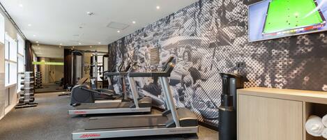 Un gimnasio con cintas de correr, dispensador de agua y una televisión mostrando un partido de billar.
