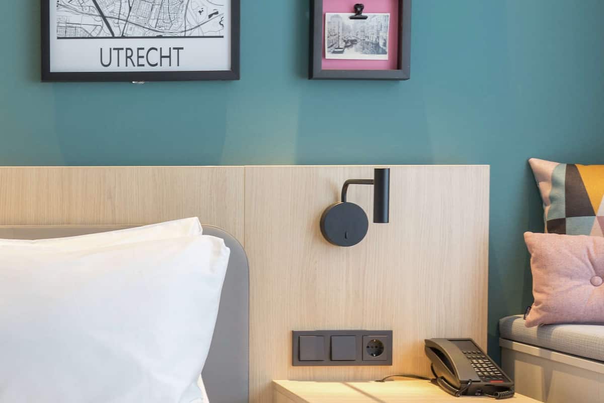 A hotel room with a bed, a nightstand with a phone and a control panel, a framed map, and a colorful abstract painting.