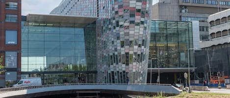A modern building with a curved glass facade, a water canal in front, and a pedestrian walkway.