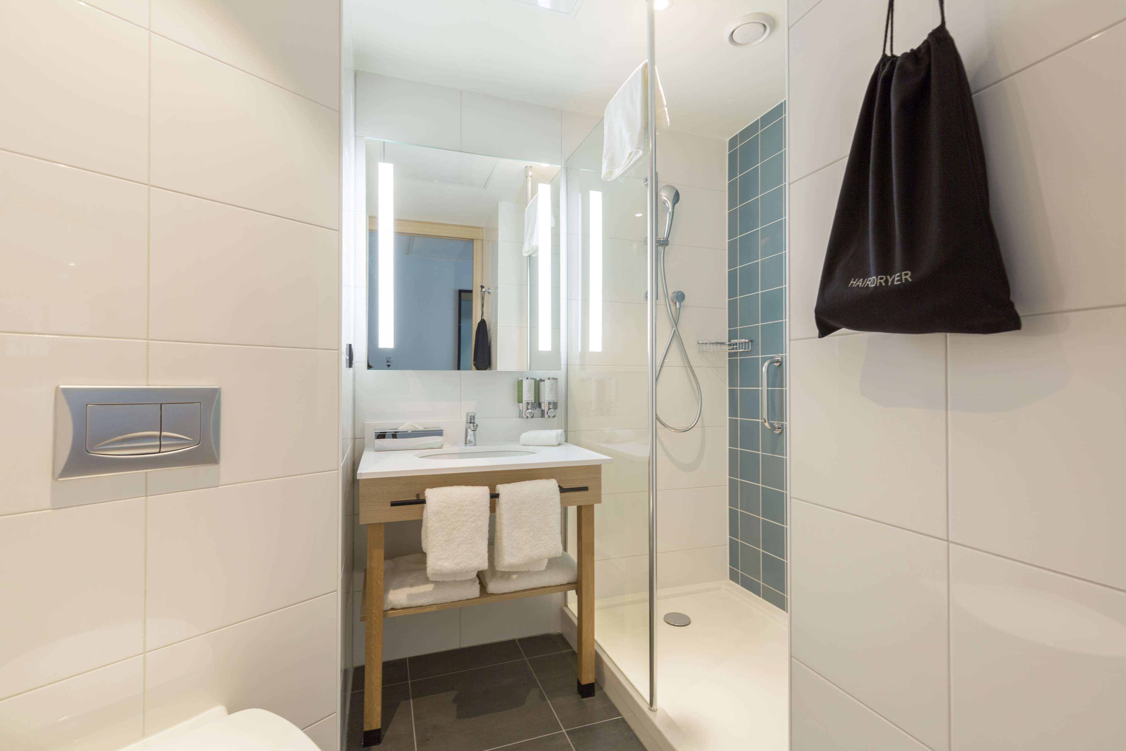 A modern bathroom with a glass shower enclosure, a wooden vanity, and a black bag hanging on the wall.