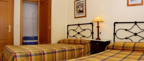 A room with two beds, a wooden door, a bathroom with a shower, and a wall-mounted lamp.