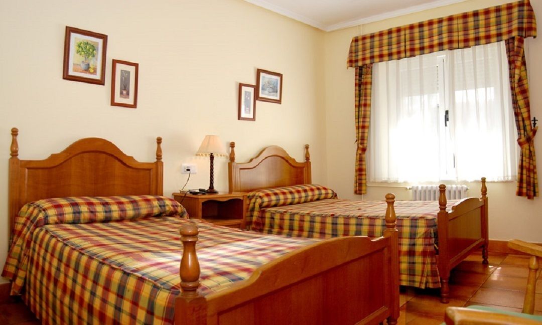 Una habitació amb dos llits de fusta, una taula de nit amb una làmpada, una finestra amb cortines i quadres emmarcados a la paret.
