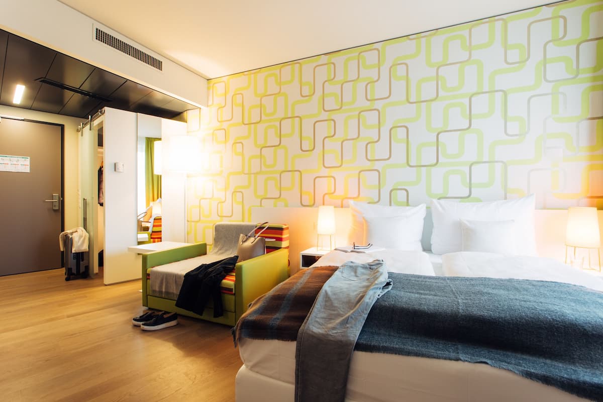 A hotel room with a bed, bedside tables, a chair, a suitcase, and a patterned wall.