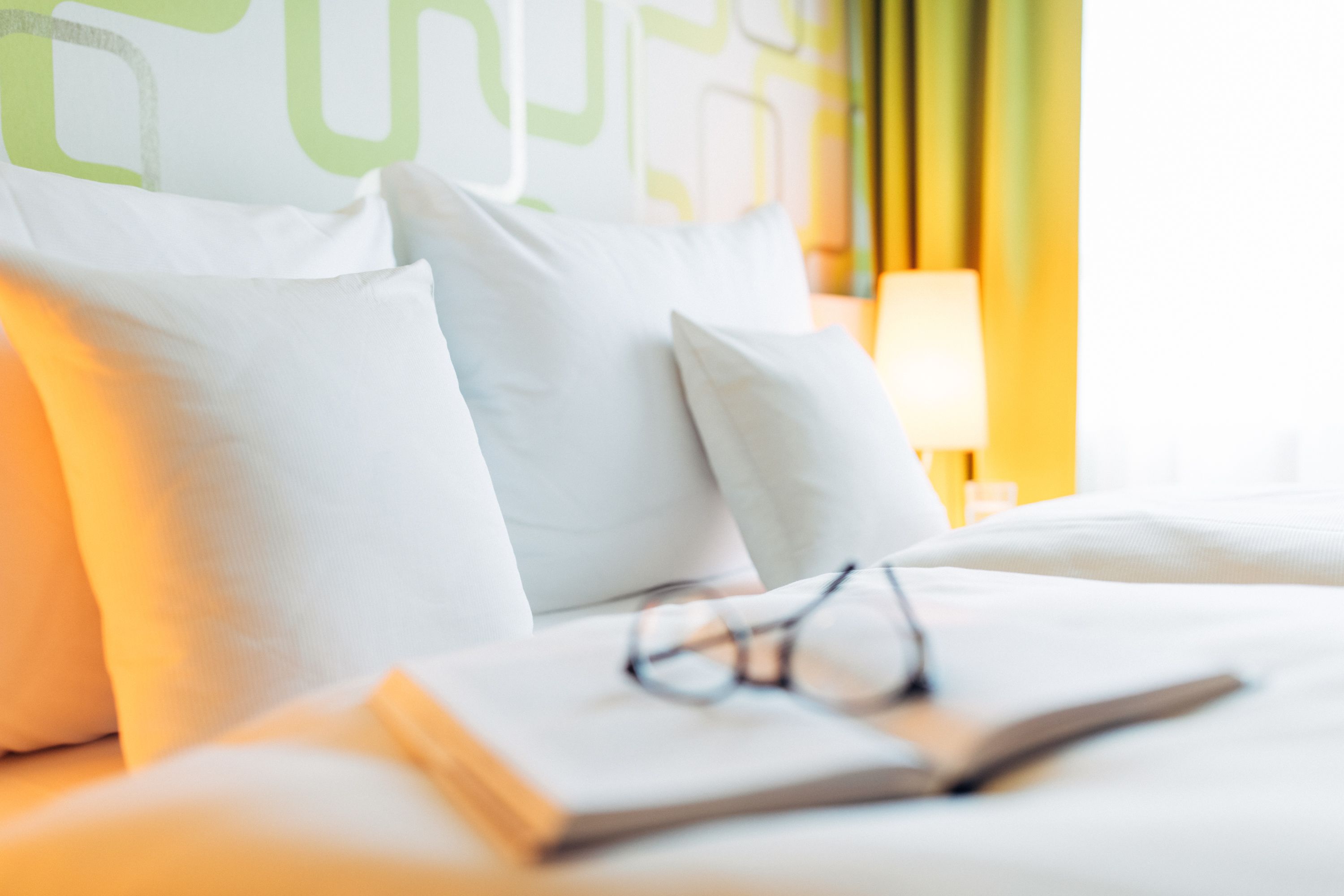 Una cama con almohadas blancas y un libro con lentes encima.