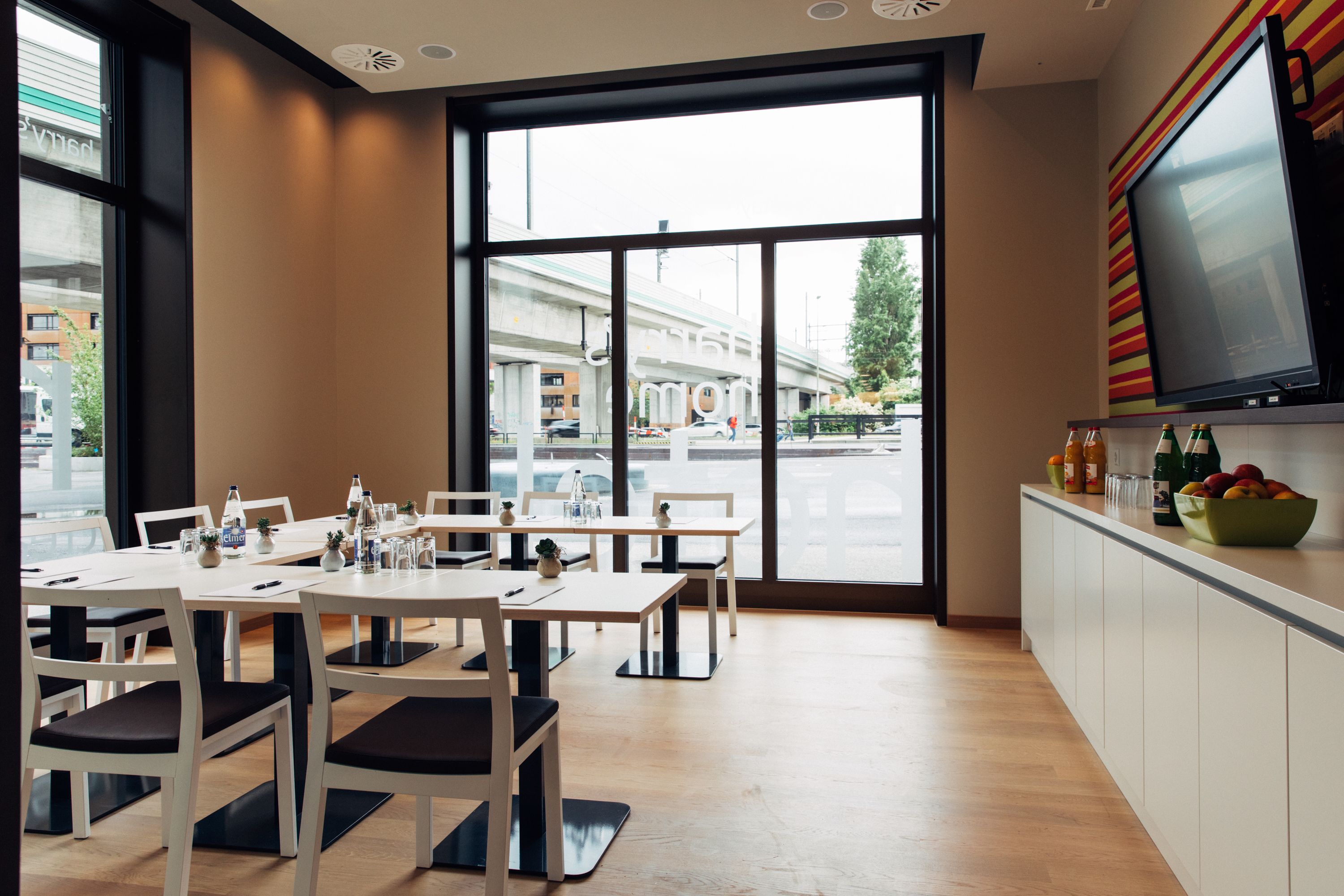 A modern dining area with large windows, white furniture, and a flat-screen TV on the wall.