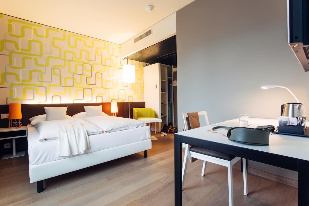 A modern hotel room with a neatly made bed, a dining table with chairs, a bright patterned wall, and a visible closet.