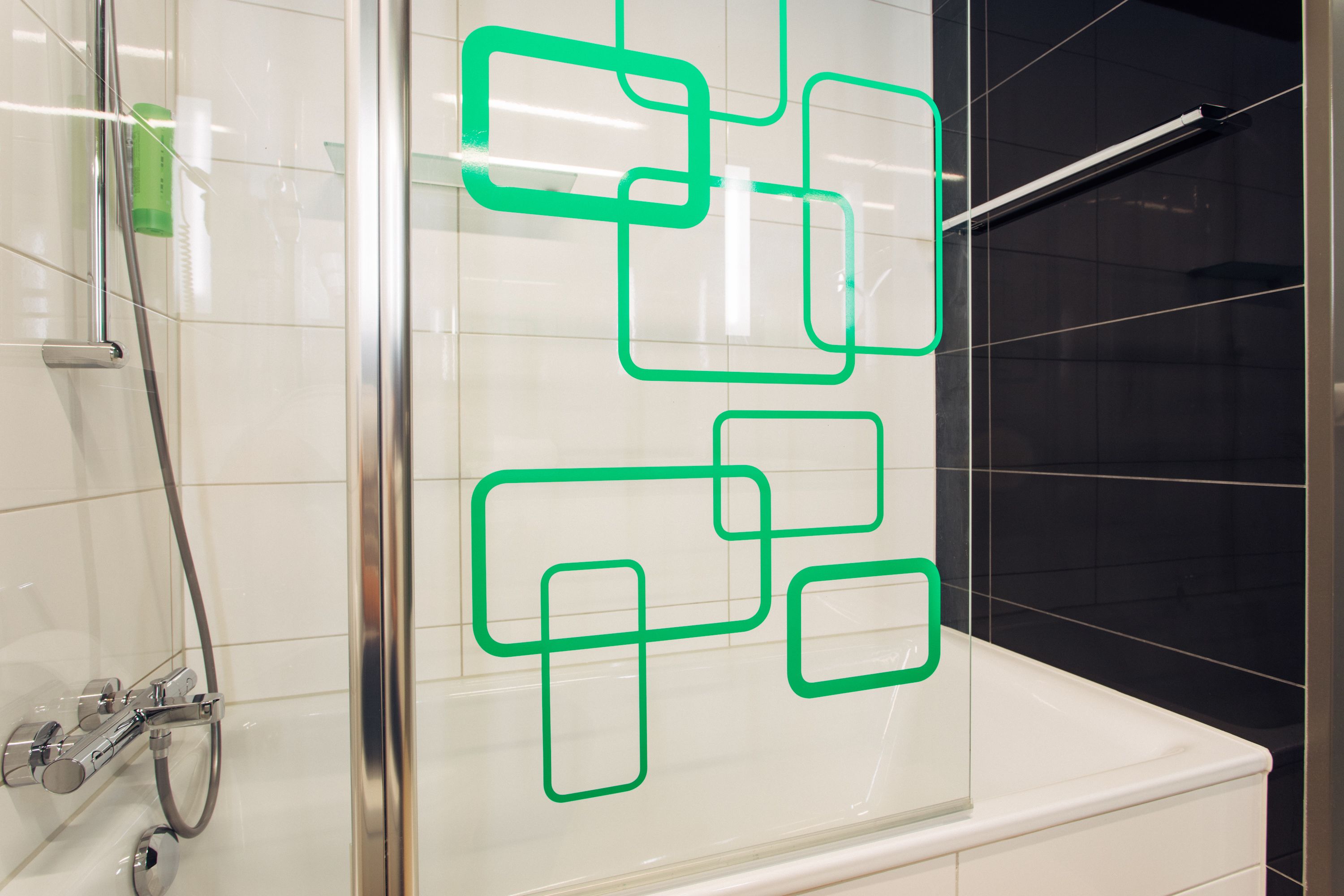 Une salle de bain avec une douche à l’italienne en verre, une baignoire blanche et un pommeau de douche vert.
