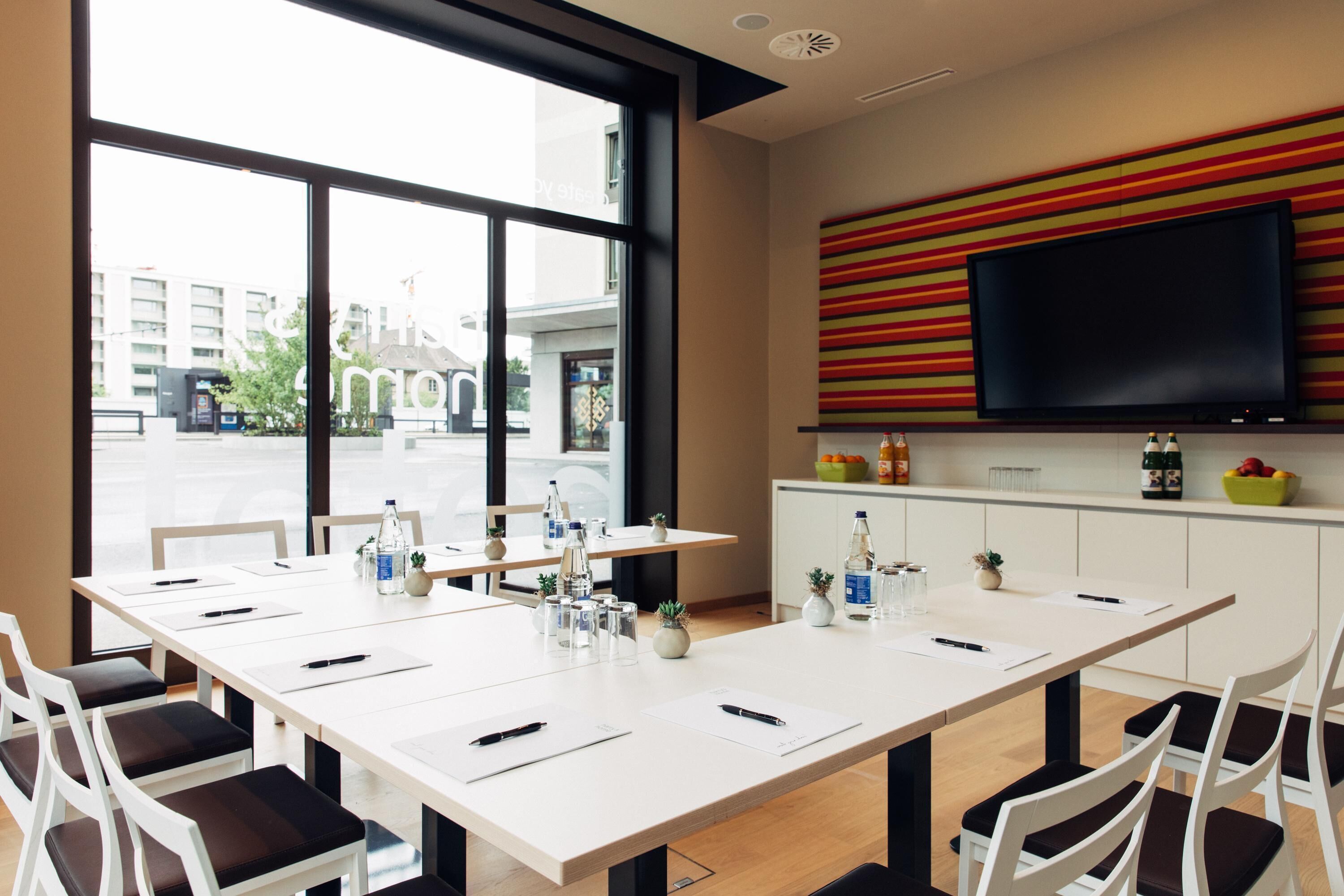 A modern conference room with a large table, white chairs, and a TV mounted on a colorful wall.