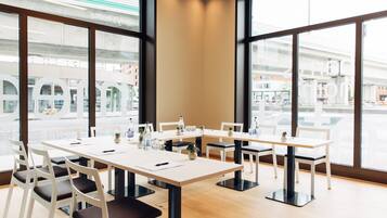 Una sala conferenze con tavoli e sedie bianche, ampie finestre e vista sulla città.