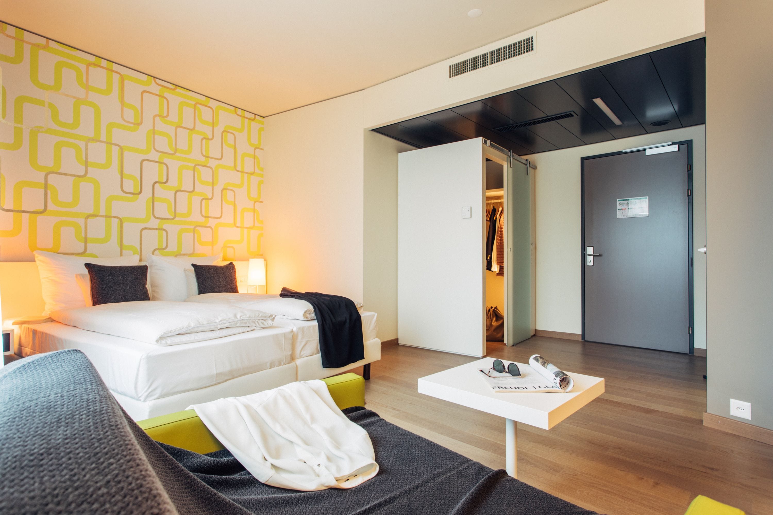 Une chambre d’hôtel moderne avec un lit soigneusement fait, une petite table sur laquelle reposent une paire de lunettes de soleil et une armoire.