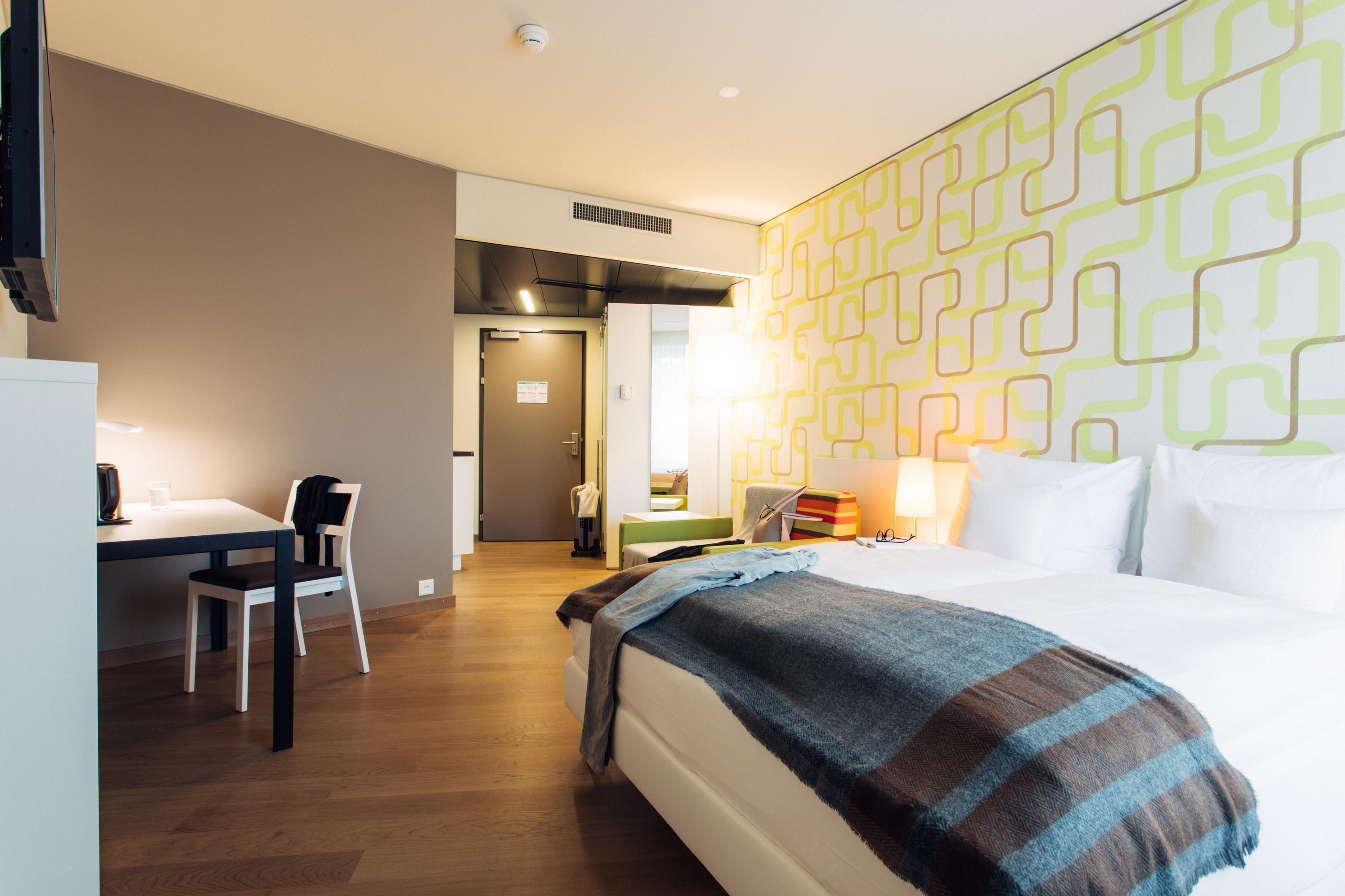 A hotel room with a bed, a desk, a chair, and a patterned wall.