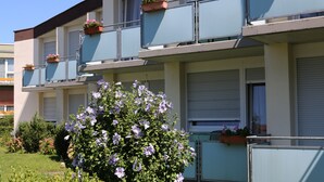 Un immeuble à plusieurs étages doté de balcons, dont certains sont ornés de jardinières fleuries, et un jardin où poussent des fleurs violettes à l'avant.