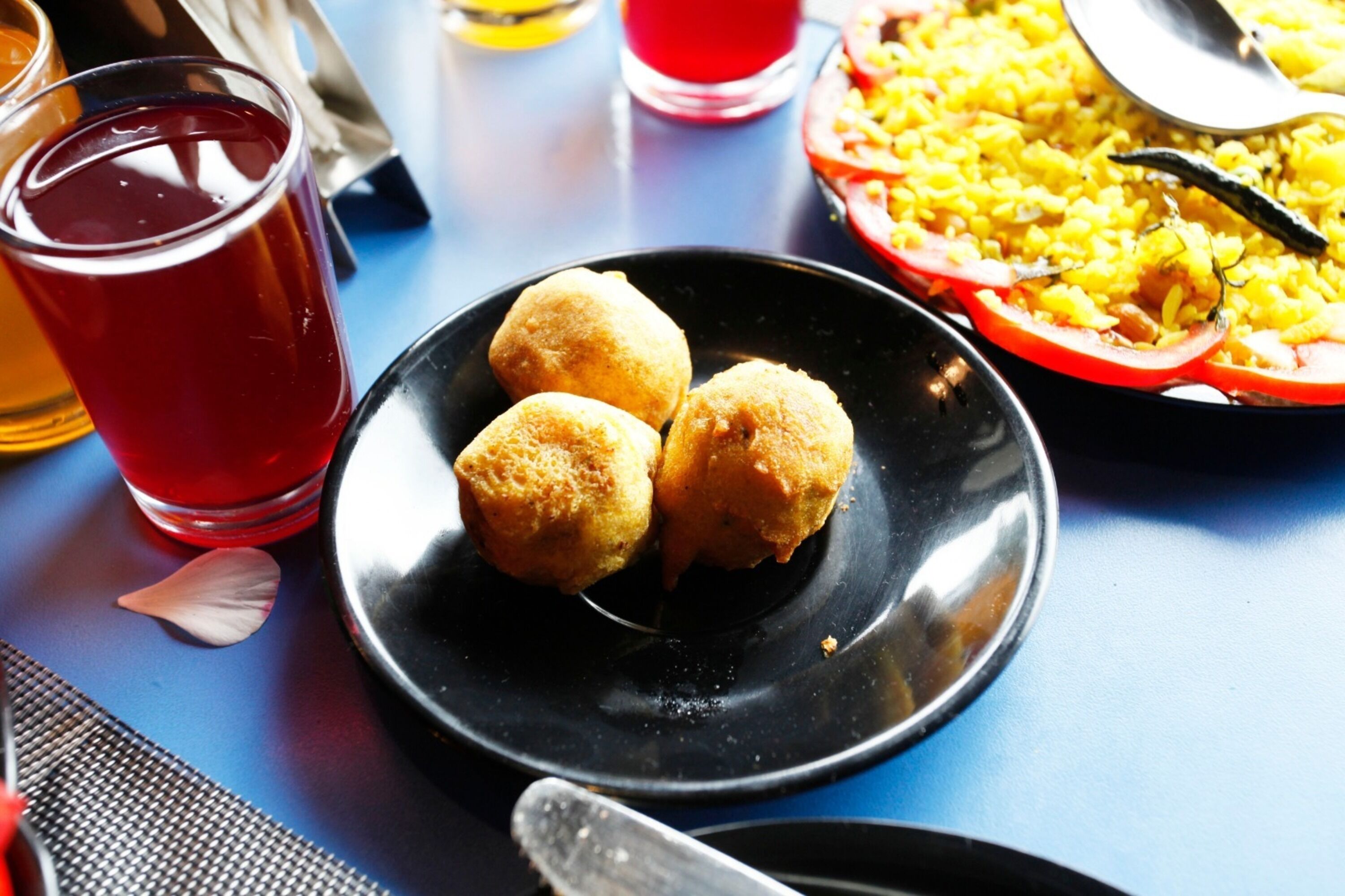 Una mesa con un plato negro que contiene tres objetos redondos, un vaso de líquido rojo y un bol de arroz amarillo con tomates.