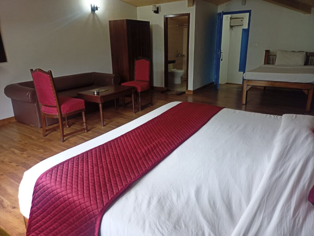 A hotel room with a large bed, a red chair, a wooden table, a sofa, and a bathroom visible through an open door.