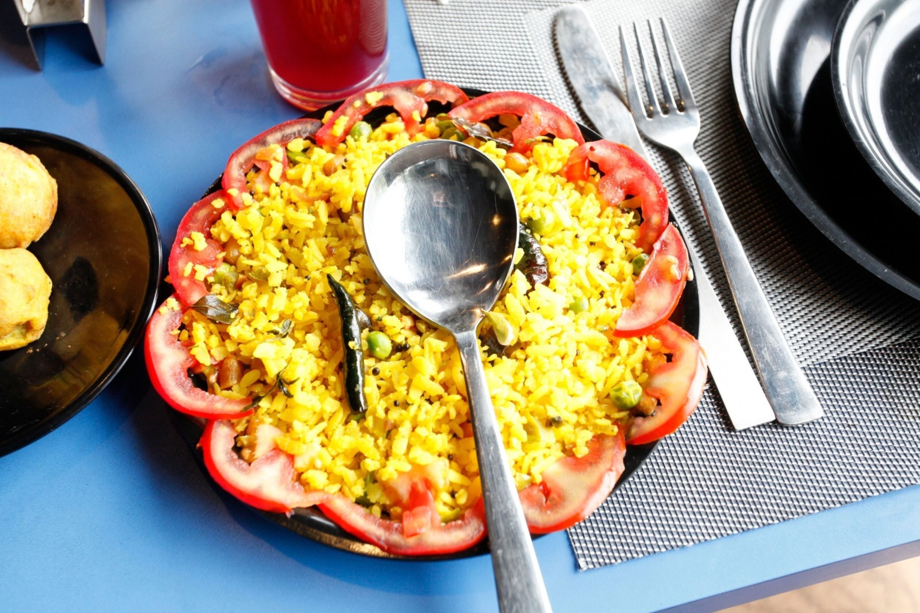A plate of yellow rice with tomato slices, a spoon, and a fork on a blue table.