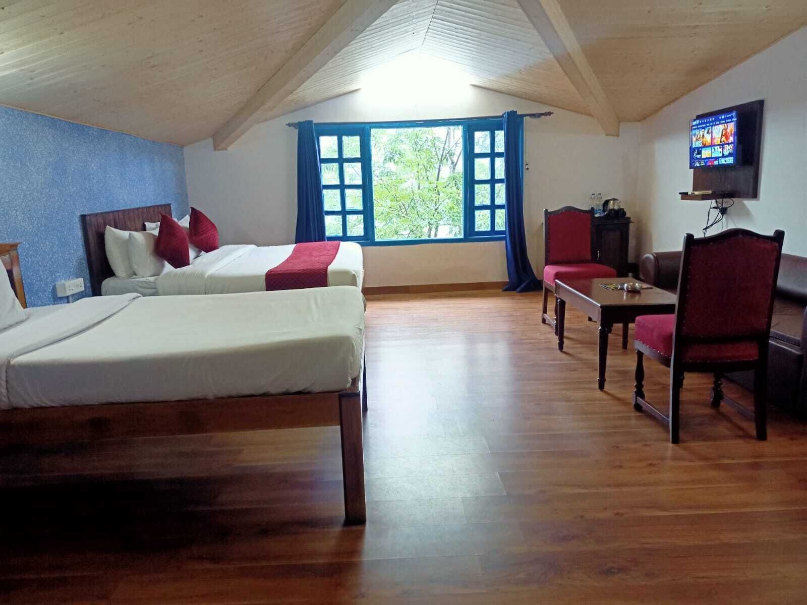 A room with two beds, a wooden floor, a TV on the wall, and a window with blue curtains.