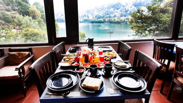 A dining table set for two with plates, glasses, and a pitcher of juice, overlooking a scenic lake and hills. - Manu Maharani Regency (Nainital)