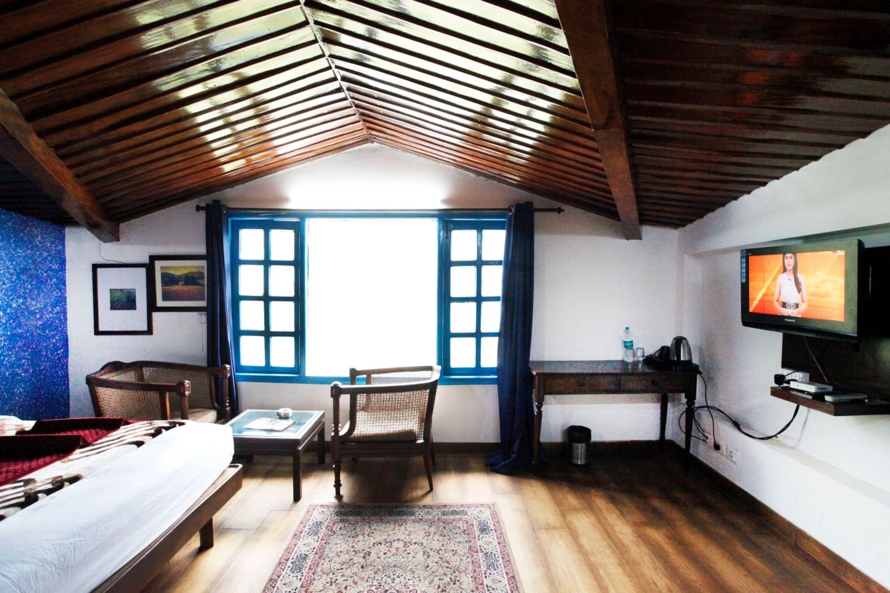 A room with a sloped ceiling, wooden beams, a bed, a desk, a chair, a television, and a window with blue curtains.