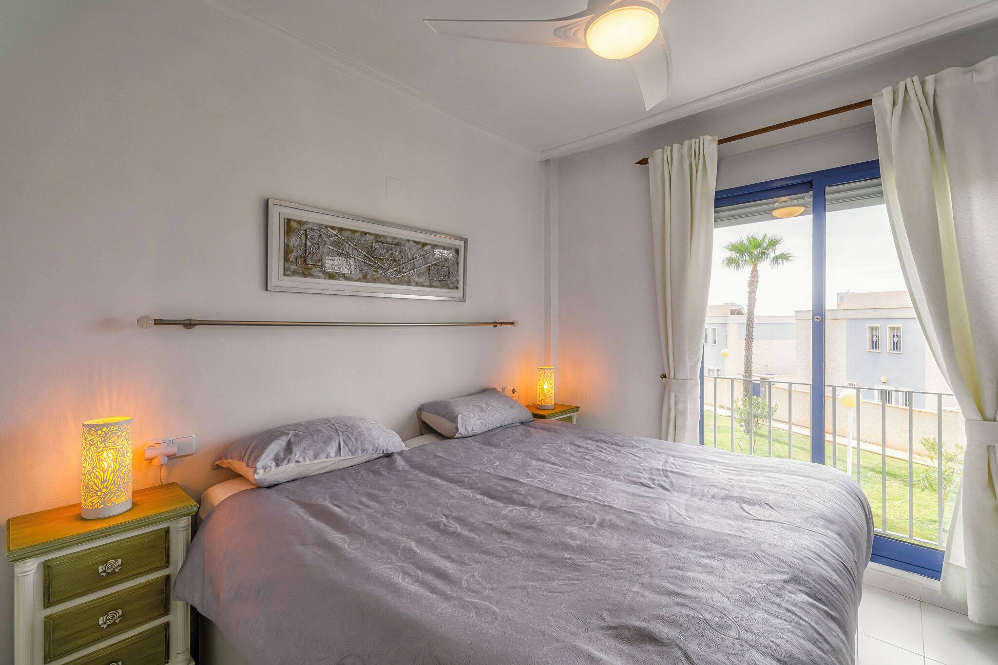 Ein Schlafzimmer mit Bett, Nachttischlampen, einem Nachttisch mit Schubladen, einem Deckenventilator, einem gerahmten Bild an der Wand und Blick auf den Balkon.