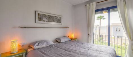 Ein Schlafzimmer mit Bett, Nachttischlampen, einem Nachttisch mit Schubladen, einem Deckenventilator, einem gerahmten Bild an der Wand und Blick auf den Balkon.