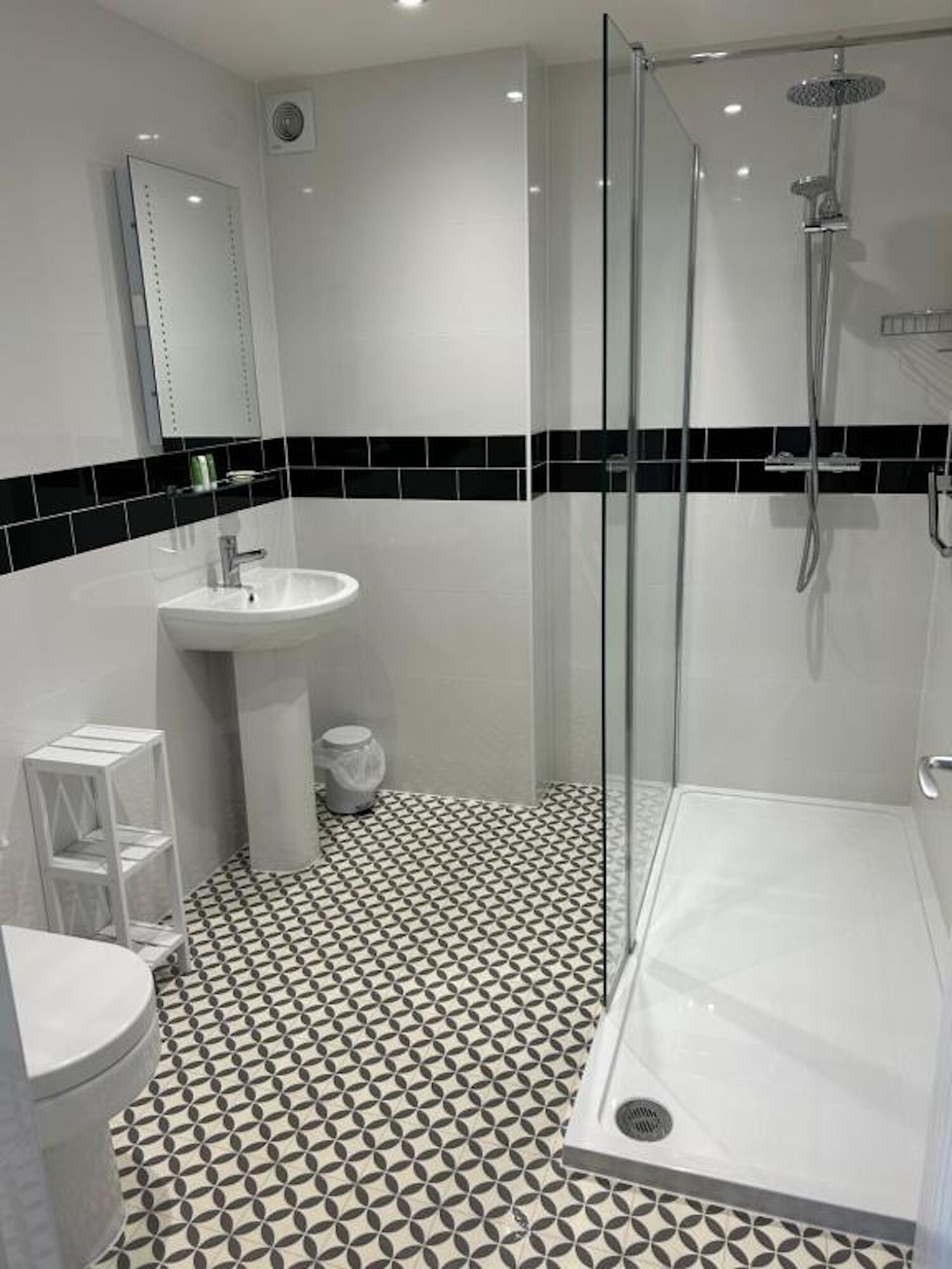 A modern bathroom with a glass shower enclosure, a white sink, and a toilet. The floor has a geometric tile pattern.
