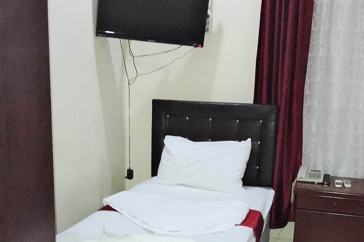 A single bed with a grey mattress and white bedding in a room with a wooden floor, a dark headboard, a flat-screen TV mounted on the wall, and a red curtain on the window.