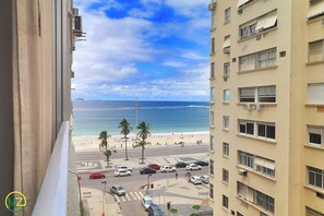 A view of a beach with a road and parked cars, a clear sky, and a building with multiple floors and balconies. - MZapartments Souza Lima (Rio de Janeiro)