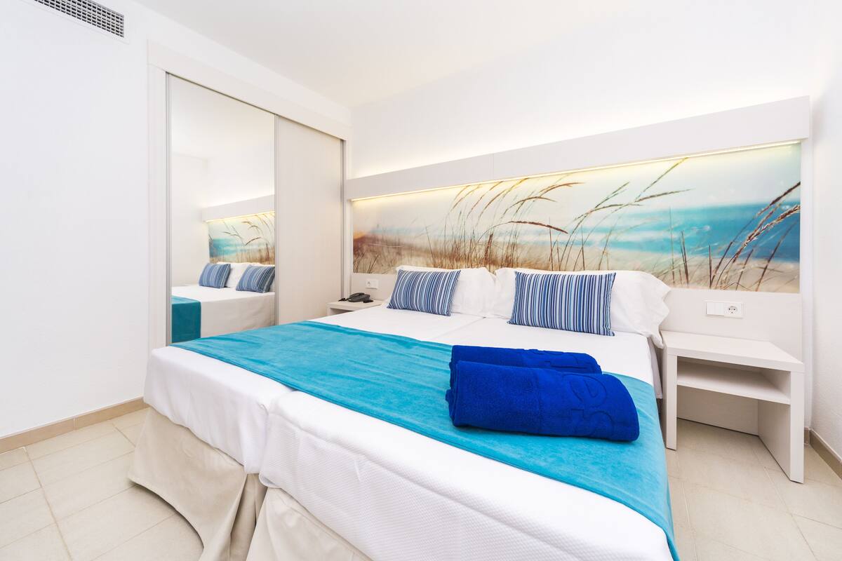 A modern hotel room with a large bed, a bedside table, a nightstand, and a large wall art depicting a beach scene.