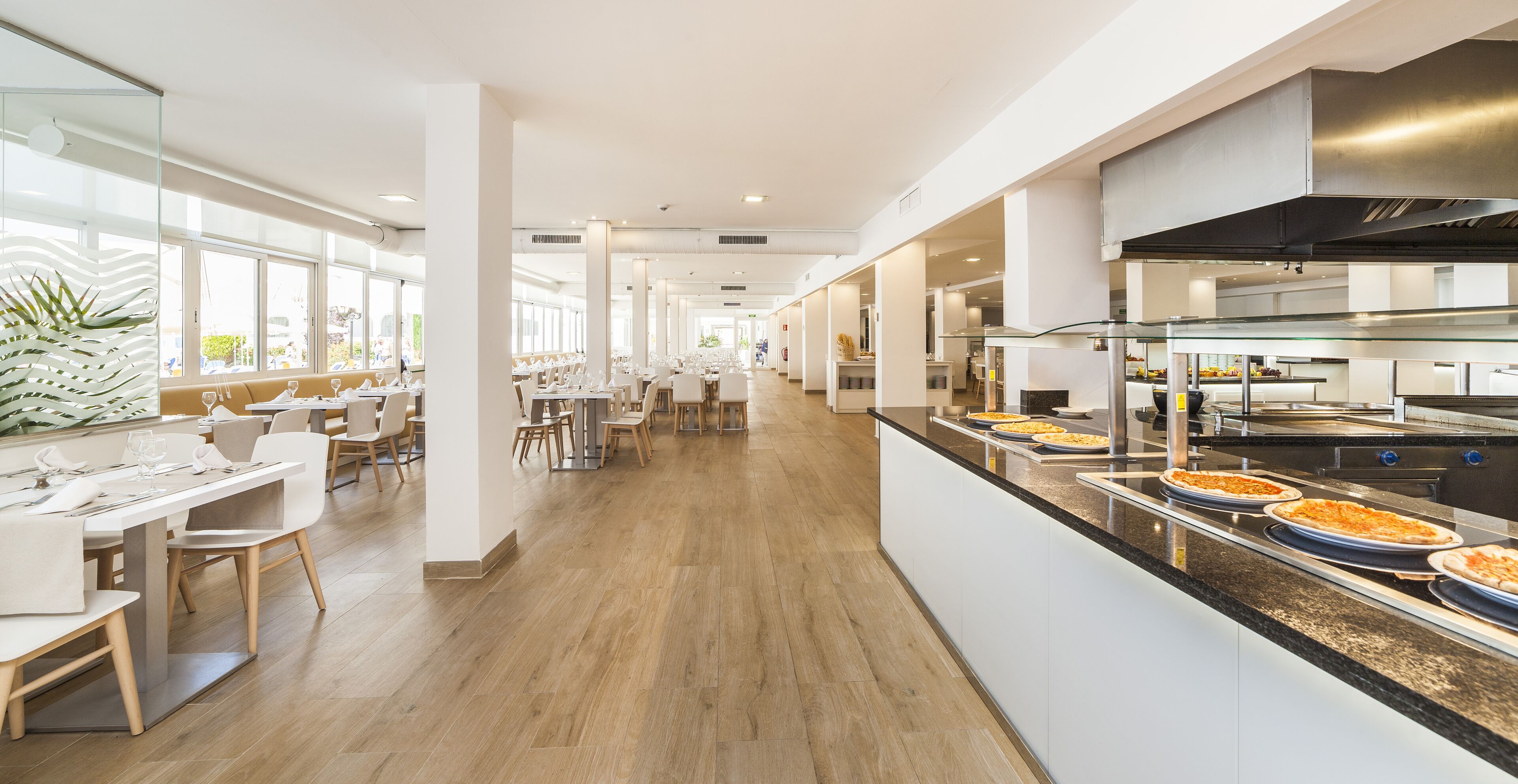 Un restaurante moderno con mesas largas, una zona de cocina y ventanas amplias.