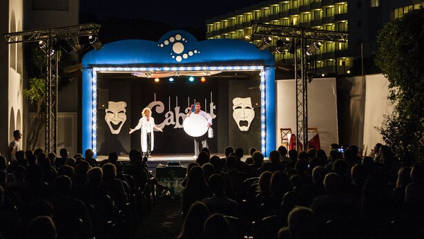 A stage with a blue backdrop and theatrical masks, surrounded by an audience in an outdoor setting. - Apartamentos Globales Lord Nelson - Adults Only (Es Migjorn Gran)