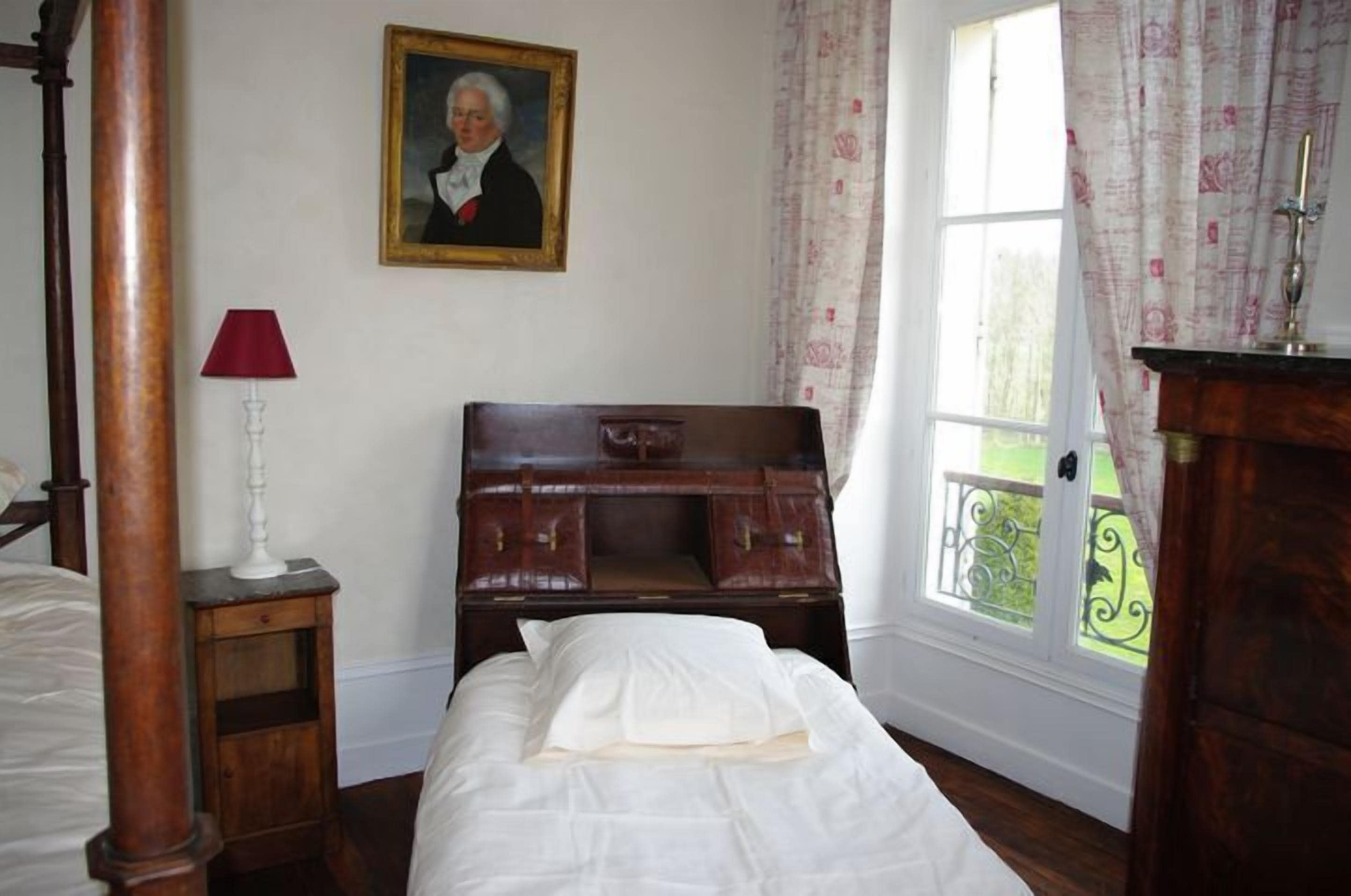 A bedroom with a wooden bed, a nightstand with a lamp, a dresser, a framed portrait on the wall, and a window with curtains.