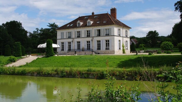 Een groot, wit, twee verdiepingen tellend huis met een symmetrische gevel, een centrale ingang en een kleine tuin met een vijver op de voorgrond.